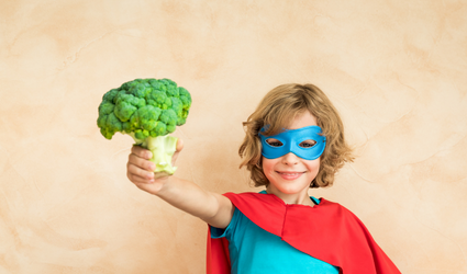 En jente utkledd som en superhelt holder opp et hode med brokkoli, som representerer sunn mat som bidrar til å forhindre ernæringsmessige mangler.