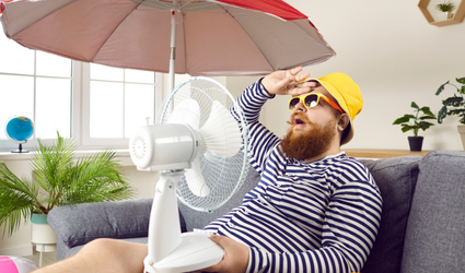En skjeggete mann med solbriller og solhatt sitter overopphetet i en sofa innendørs med en parasoll og en vifte som blåser i ansiktet.