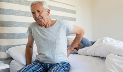 En mann med ryggsmerter har satt seg opp fra sengen og holder korsryggen med venstre hånd, tankefull, i et rom med hvite tepper og blå sengegavl.