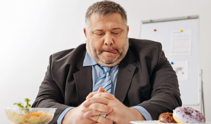 Et bilde av en overvektig mann som sitter ved et bord med sunne matalternativer på den ene siden, mens han ser lengselsfullt på smultringer mens han slikker seg om leppene.