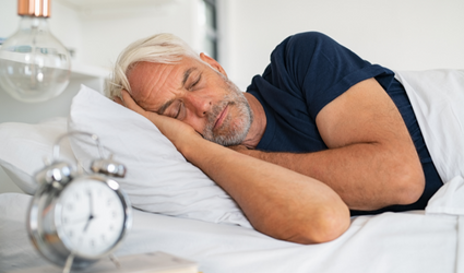 En eldre mann sover behagelig i sengen sin i morgensolen, med en klokke som viser omtrent 07:00.